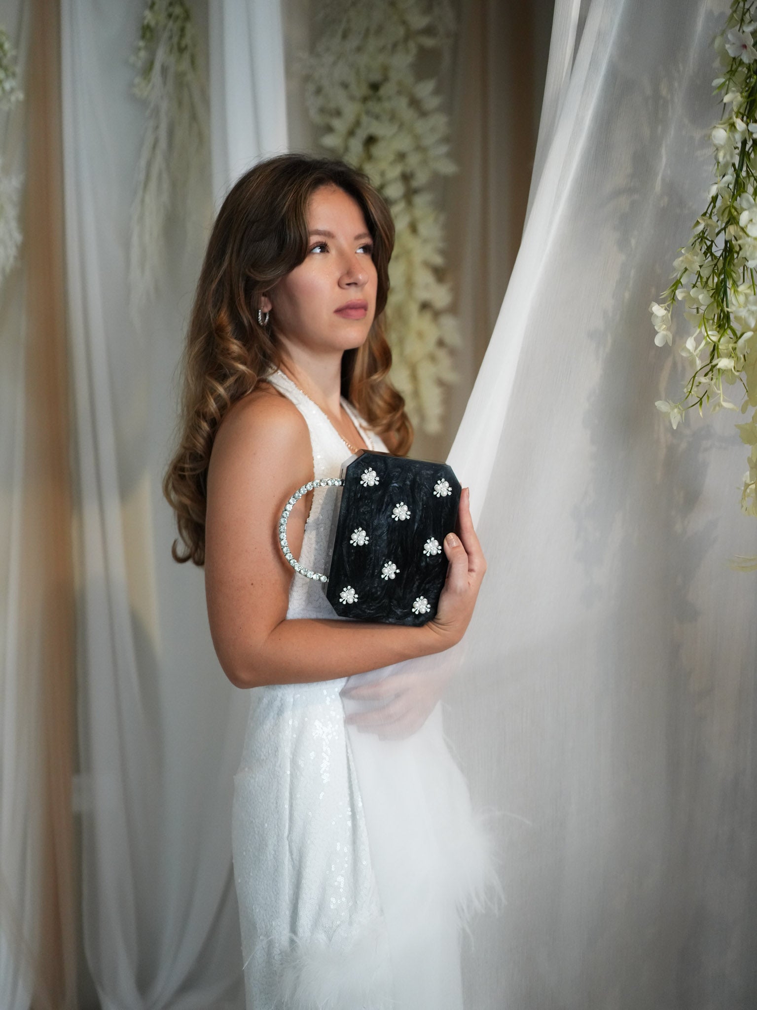 Woman in a white dress holding a black clutch with floral embellishments, standing against a floral backdrop.